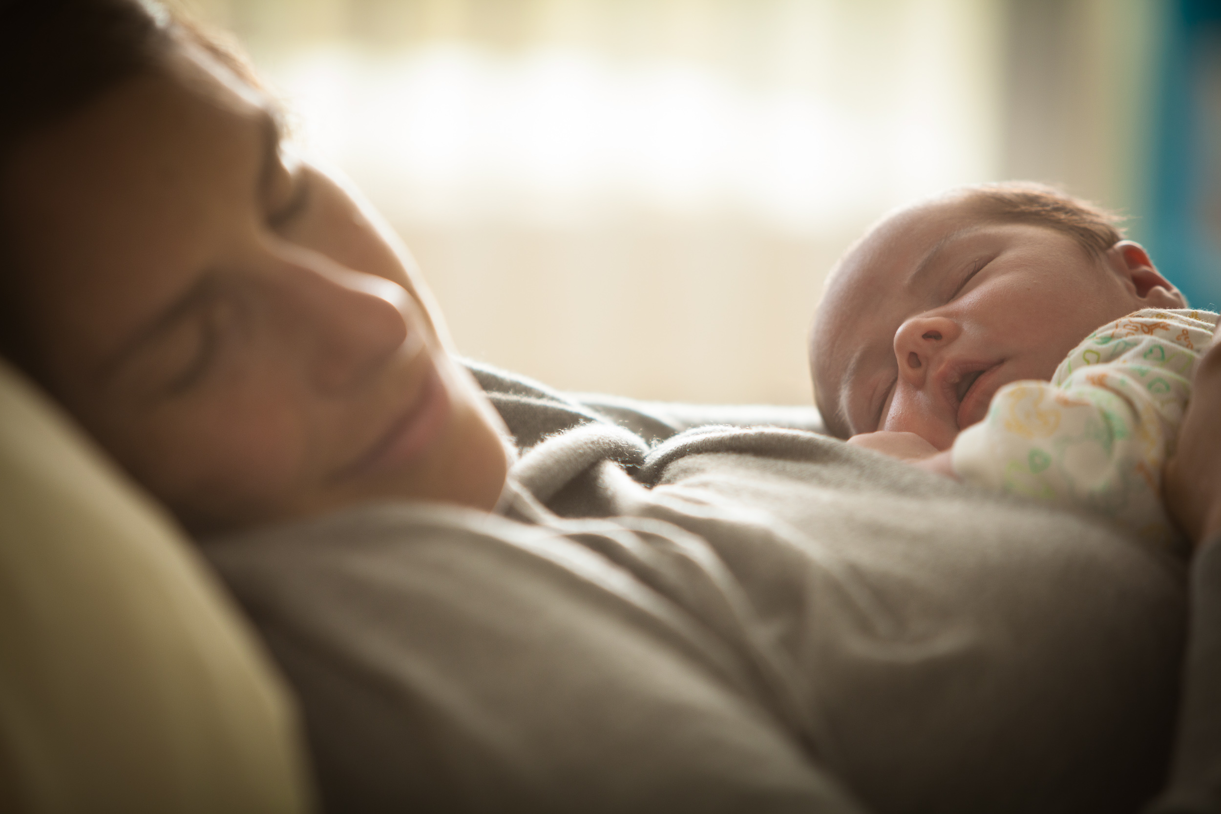 Woman and her newborn napping as the sun streams through the window of the nursery